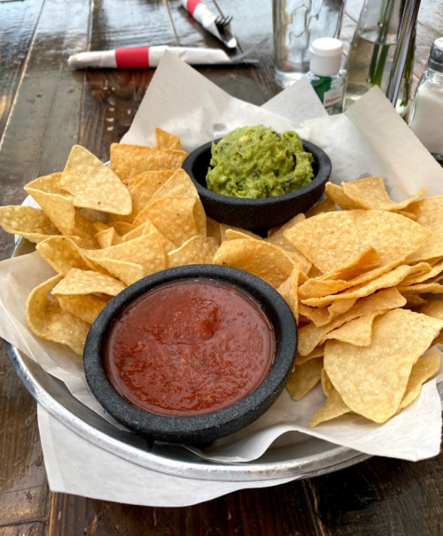 A metal bowl filled with tortilla chips, guacamole, and salsa on a wooden table.