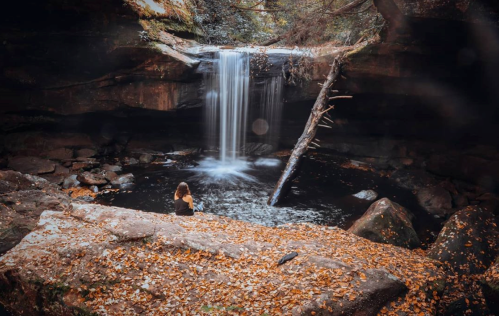 A serene waterfall cascades into a dark pool, surrounded by rocks and autumn leaves, with a person sitting nearby.