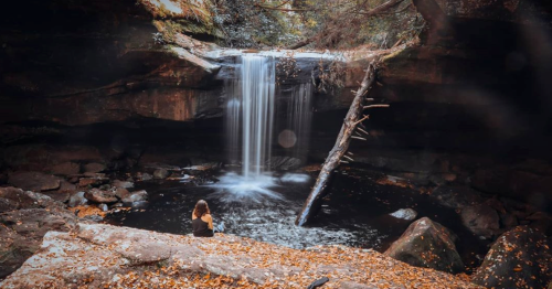 A serene waterfall cascades into a dark pool, surrounded by rocks and autumn leaves, with a person sitting nearby.