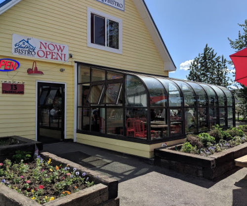 A yellow bistro with a glass-enclosed patio, colorful flowers in front, and a sign that says "Now Open!"