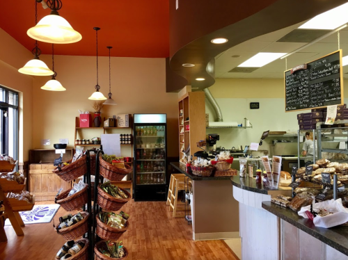 Cozy café interior with a display counter, shelves of goods, and warm lighting. Baskets filled with products are visible.