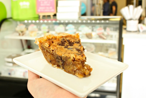 A slice of pie on a white plate, featuring a rich, gooey filling with chocolate and nuts, held in a hand.