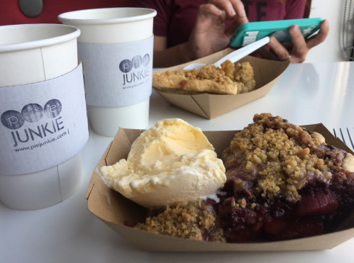 A slice of pie with ice cream and a coffee cup on a table, with a person using a phone in the background.