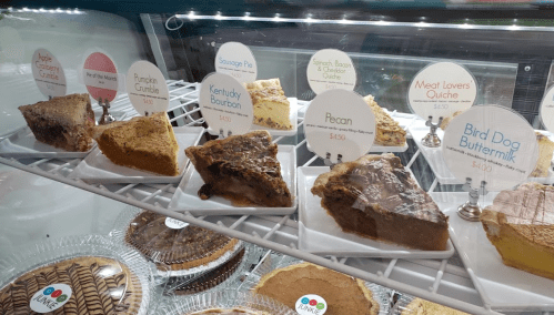 Display case filled with various desserts, including pies and quiches, each labeled with names and prices.