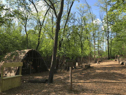 A forested area with a traditional shelter, surrounded by trees and a path, featuring a sign and natural materials.