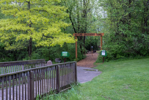 Entrance to a nature discovery trail, featuring a wooden bridge and lush greenery with a sign overhead.