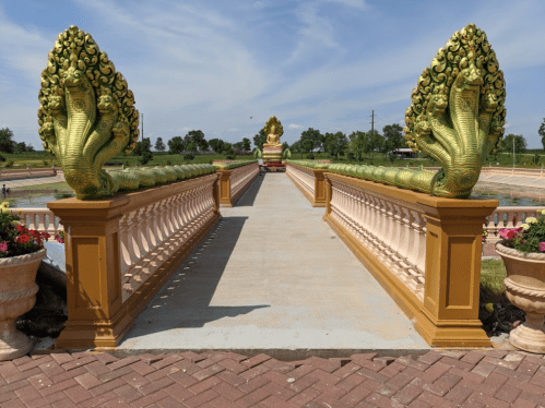 A decorative bridge with golden serpent sculptures leads to a seated Buddha statue against a blue sky.