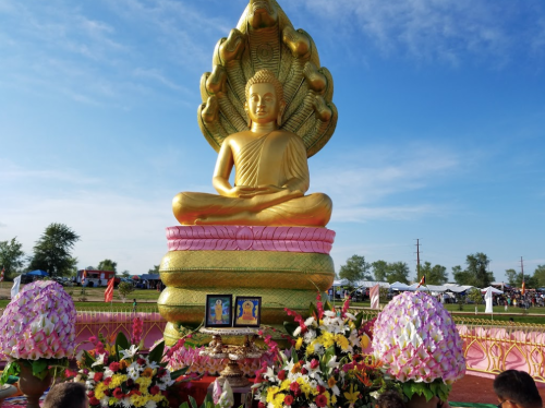 A large golden Buddha statue sits on a colorful pedestal, surrounded by flowers and offerings under a blue sky.