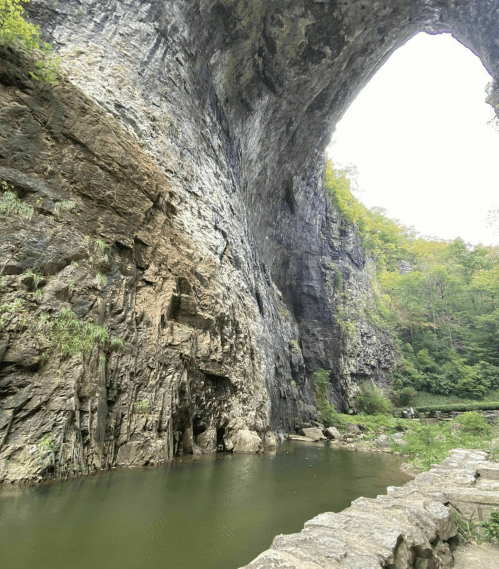 A towering rock formation arches over a calm river, surrounded by lush greenery and a serene landscape.