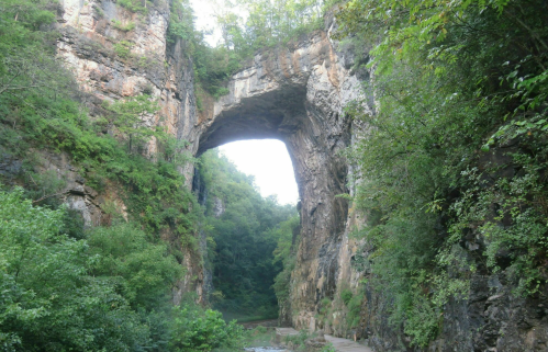 Natural stone arch surrounded by lush greenery and rocky cliffs, with a pathway leading through the landscape.