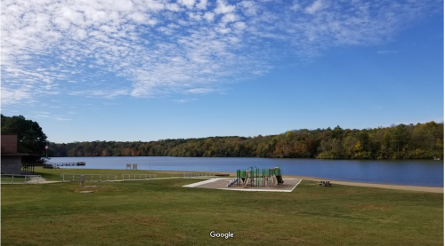 A serene lake view with a playground and grassy area, surrounded by trees and a blue sky with scattered clouds.