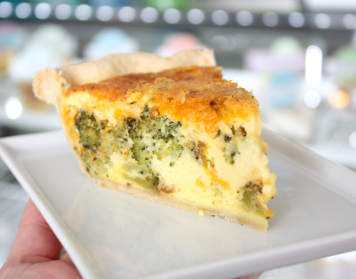 A slice of broccoli cheese quiche on a white plate, showcasing a golden crust and creamy filling with green broccoli pieces.