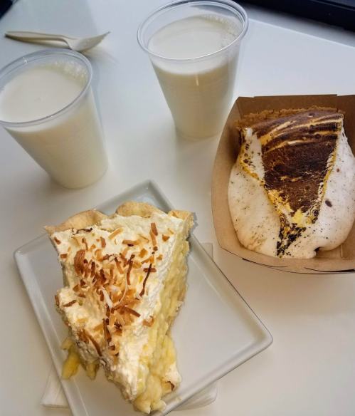 Two cups of milk beside a slice of coconut cream pie and a s'mores dessert in a takeout box on a white table.