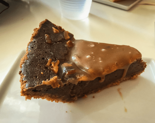 A slice of chocolate cake with a glossy caramel topping and bits of nuts on a white plate.