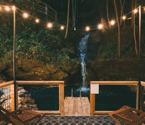 A cozy wooden deck with string lights overlooks a serene waterfall and dark pool surrounded by lush greenery.