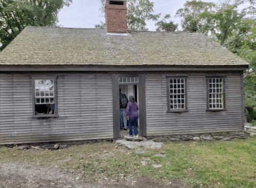 A rustic wooden house with a chimney, featuring a person entering through the front door. Trees are visible in the background.