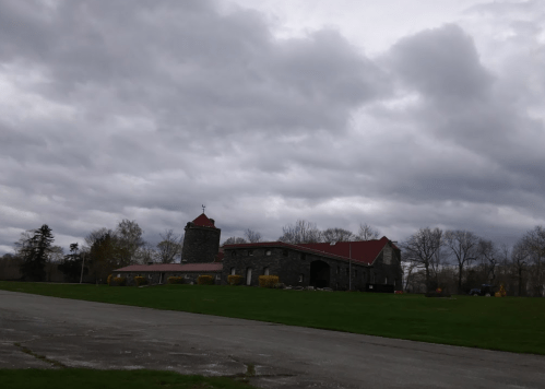 A stone building with a red roof sits on a grassy field under a cloudy sky. Trees are visible in the background.