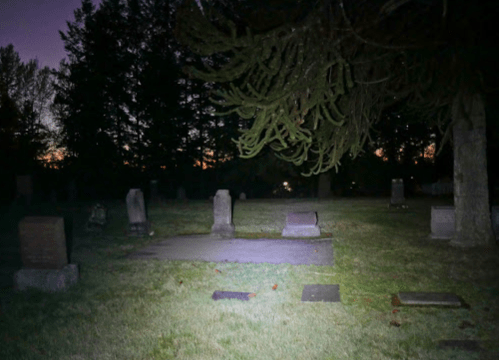 A dimly lit cemetery at dusk, featuring gravestones and trees silhouetted against a fading sky.