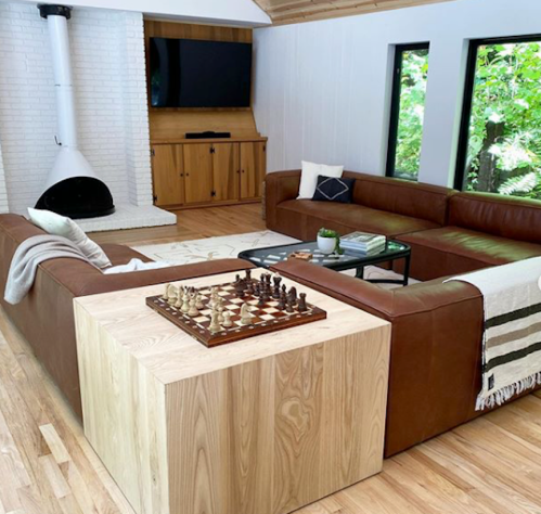 A modern living room with a leather sectional, coffee table, chess set, and large windows overlooking greenery.