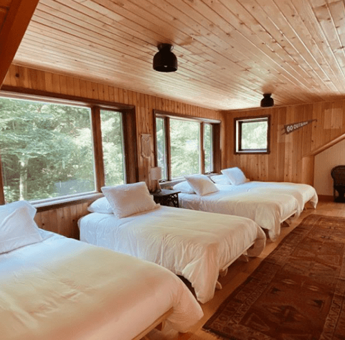 Cozy wooden bedroom with three beds, large windows, and warm lighting, surrounded by nature.