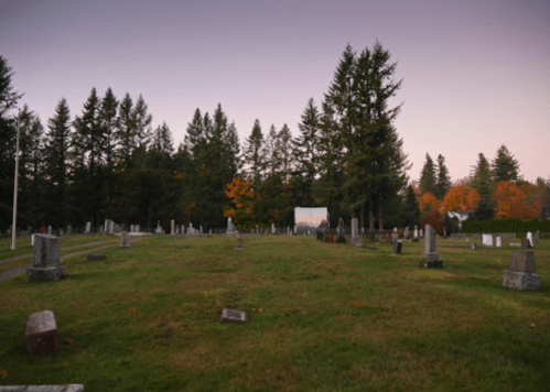 A serene cemetery at dusk, surrounded by tall trees and colorful autumn foliage.