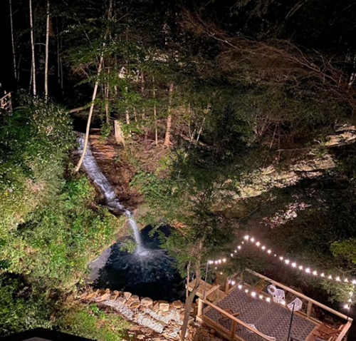 A serene nighttime scene of a flowing stream surrounded by trees, with a deck illuminated by string lights.