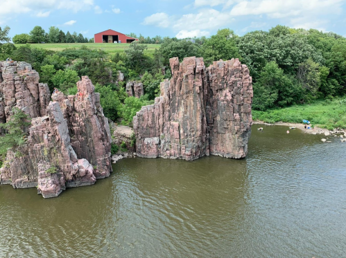 Rocky cliffs rise above a calm river, with lush greenery and a red barn in the background under a blue sky.