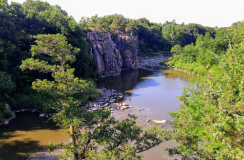 A serene river flows past rocky cliffs, surrounded by lush green trees under a clear blue sky.