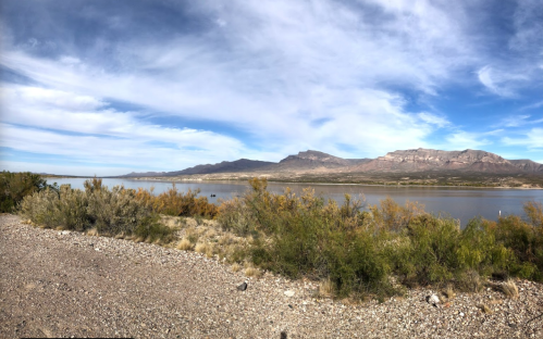 Panoramic view of a calm lake surrounded by mountains and lush vegetation under a blue sky with scattered clouds.