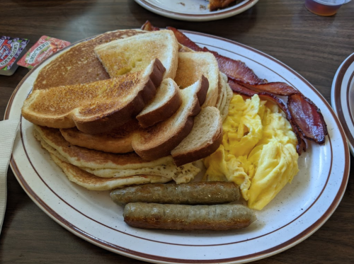 A plate of breakfast featuring pancakes, toast, scrambled eggs, sausage links, and crispy bacon.