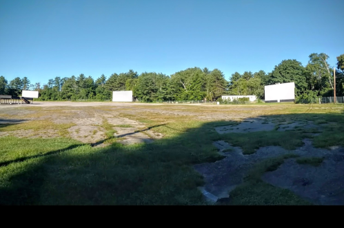 A wide view of an empty drive-in theater with two large screens and a grassy area under a clear blue sky.