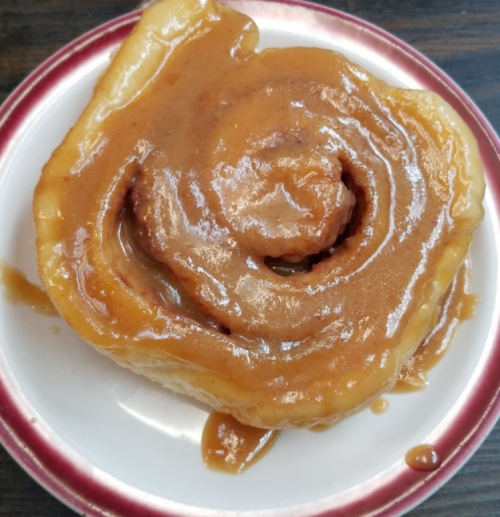 A close-up of a sticky cinnamon roll topped with a glossy caramel glaze on a white plate with a red rim.