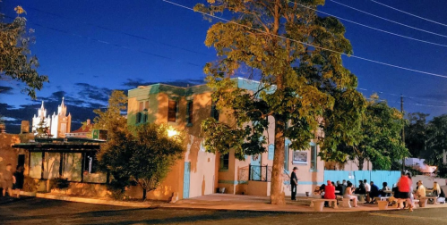 A lively outdoor scene at dusk, featuring a building with turquoise accents and people gathered nearby under trees.