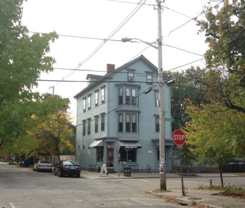 A three-story blue building at a street corner, surrounded by trees and parked cars, with a stop sign nearby.