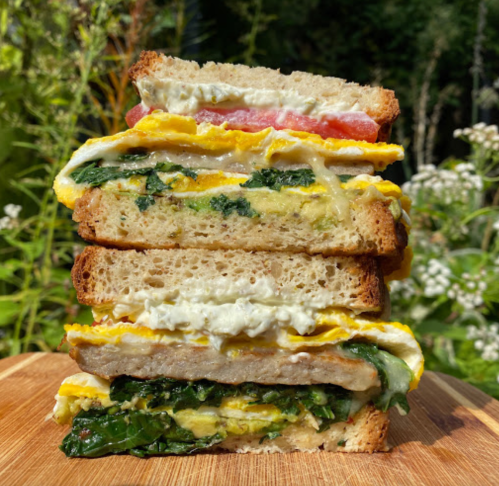 A tall sandwich stacked with layers of eggs, spinach, tomato, and creamy spread, resting on a wooden board outdoors.