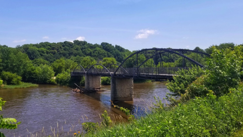 A historic bridge spans a river, surrounded by lush greenery and rolling hills under a clear blue sky.
