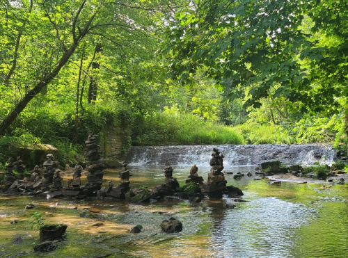 A serene stream with stacked stones and a gentle waterfall, surrounded by lush green trees and foliage.