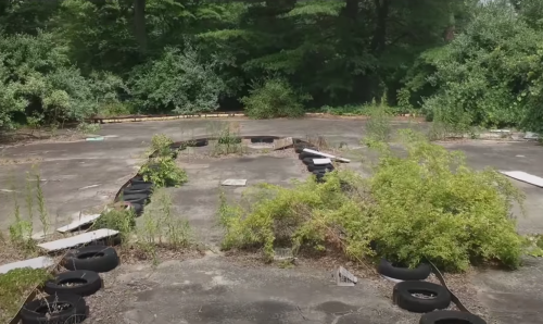 Overgrown, abandoned area with cracked concrete, scattered tires, and vegetation reclaiming the space.