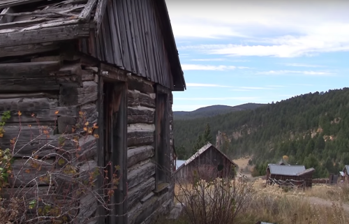 A weathered wooden cabin in a mountainous landscape, surrounded by trees and distant hills under a blue sky.