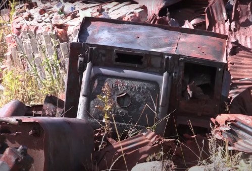 Rusty, abandoned machinery and metal debris scattered among overgrown grass and rubble.