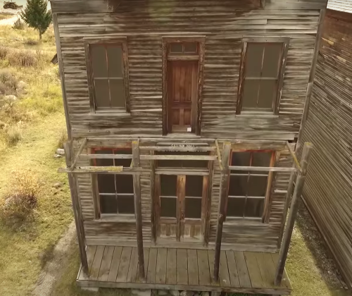 A weathered, two-story wooden building with a porch, surrounded by grassy terrain and other old structures.