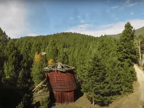 A weathered, rusted silo stands among tall green trees, with a dirt path winding through the forest in the background.