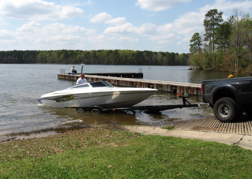 A man launches a white boat from a trailer into a lake, with a dock and trees in the background.