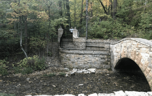 A stone bridge arches over a small stream, surrounded by trees with autumn foliage. A person stands on the bridge.