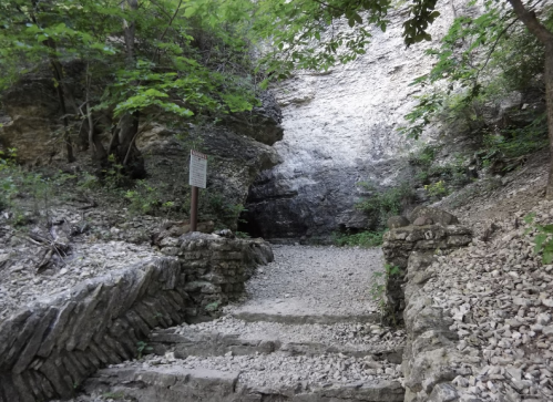 Stone steps lead up to a rocky entrance surrounded by lush greenery in a natural setting.