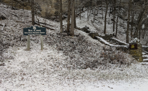 Sign for Ice Cave in a snowy forest, with stone ruins visible nearby among the trees.