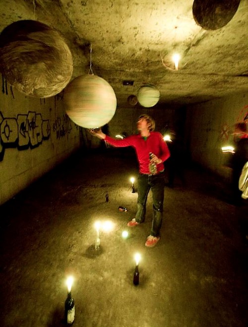 A person in a red shirt interacts with hanging spheres in a dimly lit underground space surrounded by candles.