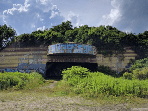 Abandoned structure covered in graffiti and overgrown vegetation, set against a cloudy sky.