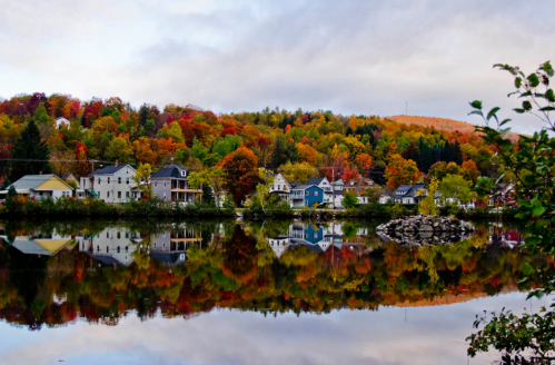A serene lake reflects colorful autumn foliage and charming houses along the shore, under a cloudy sky.