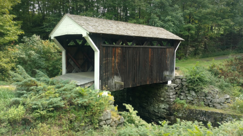 A rustic covered bridge made of wood, resting on stone supports, surrounded by lush greenery and trees.